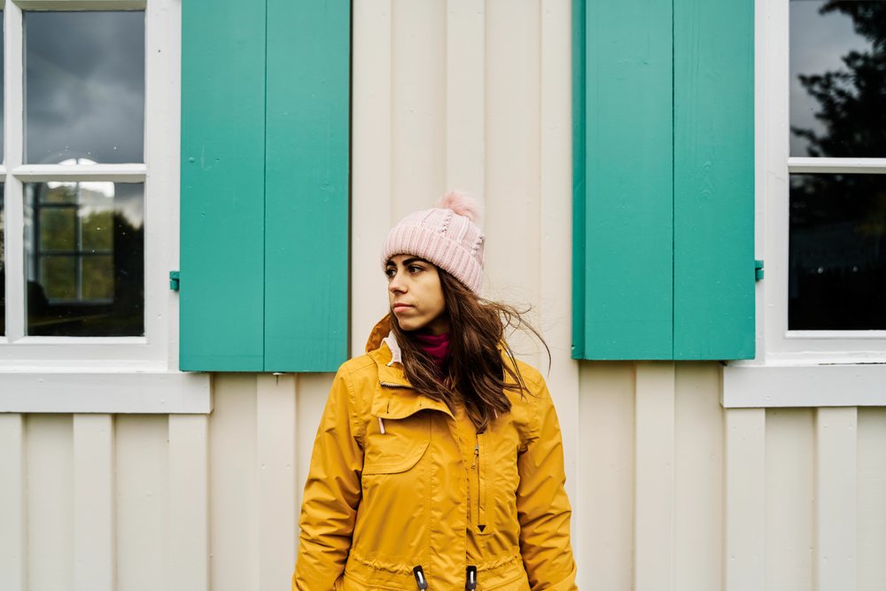 A woman wearing a yellow jacket and pink knit hat stands outdoors against a cream-colored wall with green window shutters, Iceland.