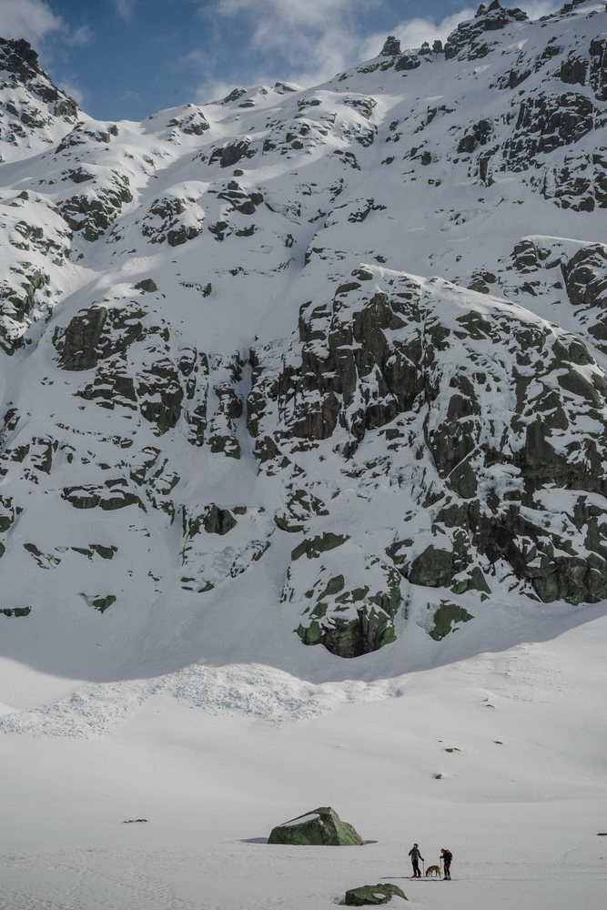 Two people snowshoeing with a dog near large rocks under snow-covered mountains, mountaineering, climbing, landscape, Spain.