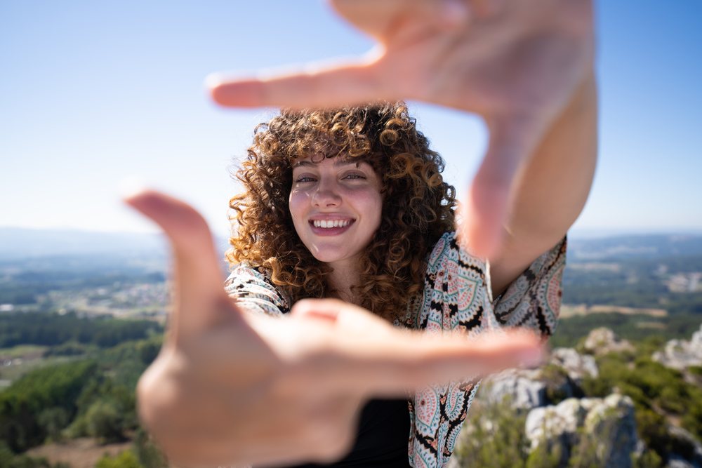 Smiling woman with curly hair framing her hands outdoors with mountainous landscape and green forest under a clear blue sky, Pico Sacro, Galicia, Spain.
