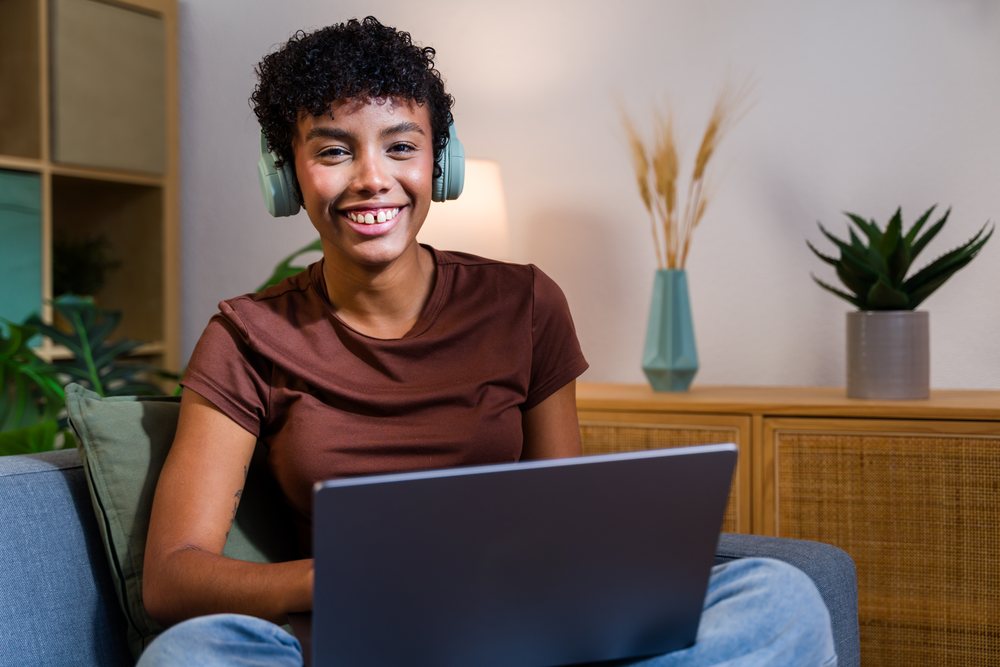Smiling woman wearing headphones uses a laptop while seated on a sofa in a modern living room with indoor plants and wooden cabinet, Spain.