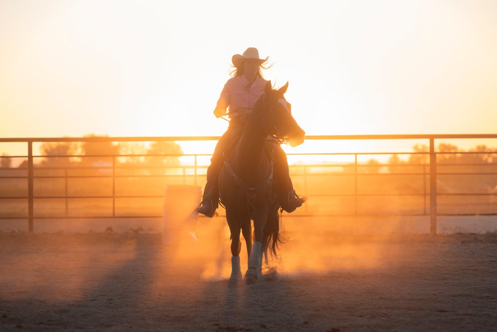 Horseback rider wearing a cowboy hat moves through a dusty fenced arena at sunset, Southwest USA.