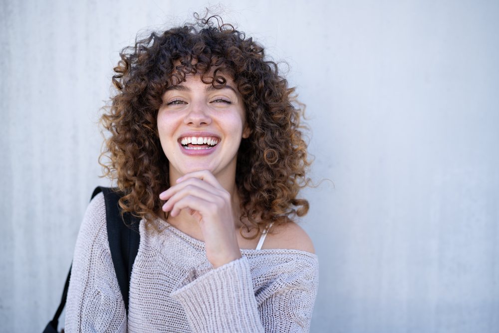Young woman with curly hair smiling happily outdoors against a plain wall, home story, influencer, Spain.