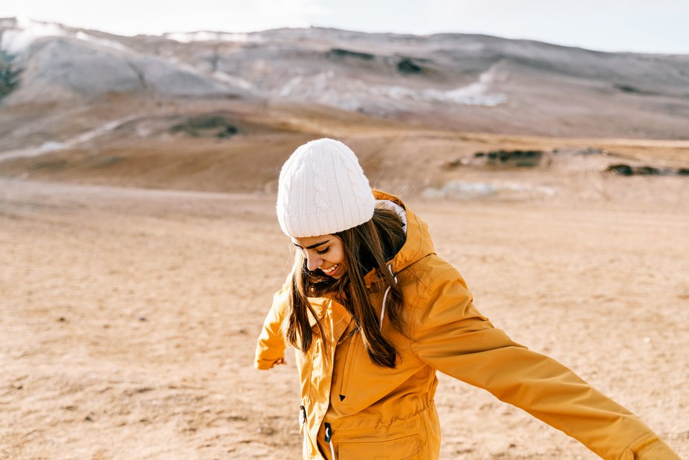 A woman wearing a yellow jacket and white beanie stretches her arms outdoors in a brown mountainous landscape, Iceland.