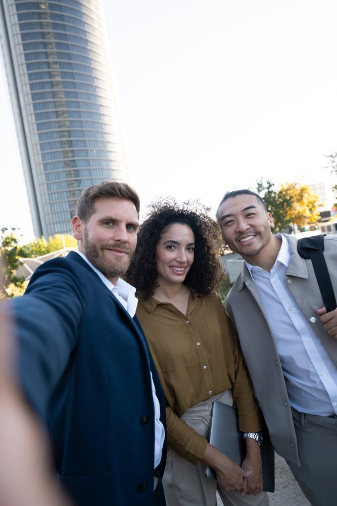 Three business professionals smile while taking a selfie outdoors by a modern building, Madrid, Spain.