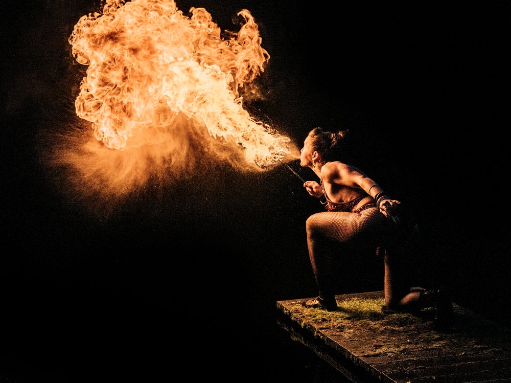 Woman kneeling on a mossy platform breathes fire with dramatic lighting against a dark background, artistry, artist, USA.
