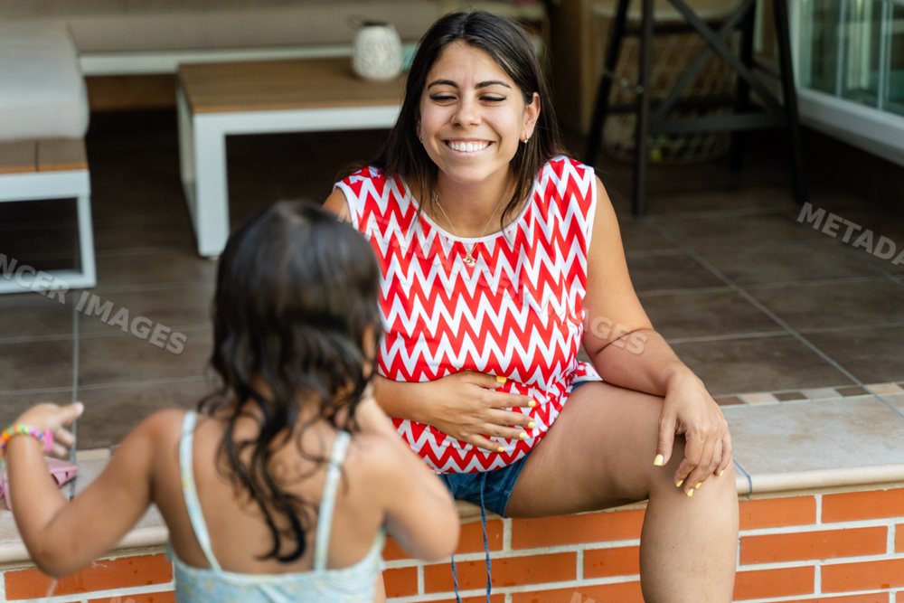 A smiling white woman sits on a brick ledge interacting with a child