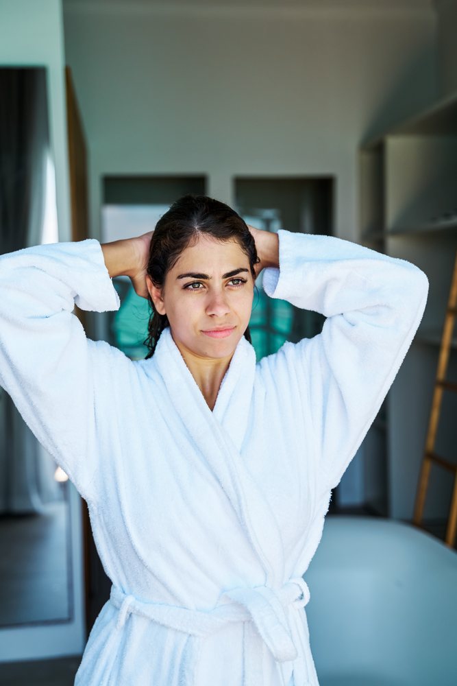 A woman wearing a white bathrobe stands indoors with her hands behind her head in a bathroom setting in the, Maldives.
