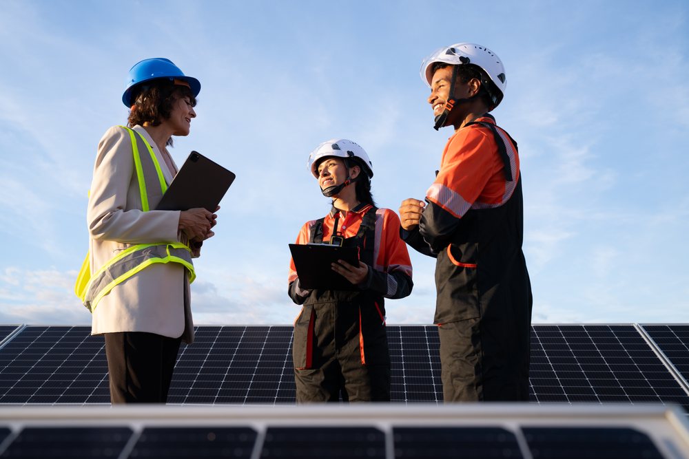 Three engineers in helmets and safety vests inspect solar panels outdoors under a sunny blue sky, Madrid, Spain.