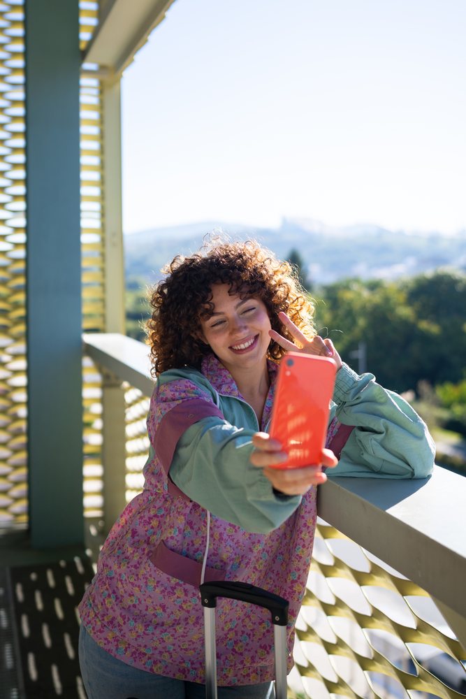 Woman with curly hair takes a selfie making a peace sign on a balcony overlooking greenery, Spain.