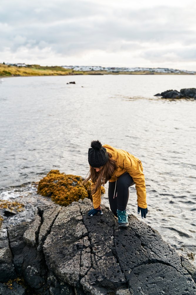 A white woman climbs volcanic rocks along a rocky shore