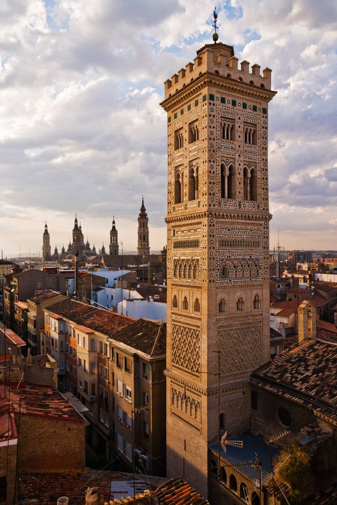 Shrines of the Two Marys tower rises above rooftops at sunset under a partly cloudy sky, Zaragoza, Spain.