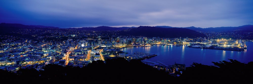 Nighttime cityscape of Wellington, New Zealand showing illuminated harbor and skyline reflecting on water with hills and mountains in view.