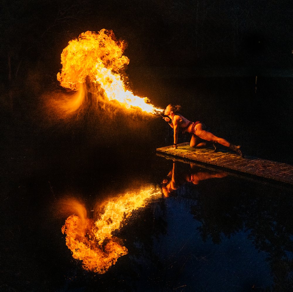 Woman performing fire breathing on a dock at night with large flames reflected in the water, artistry, artist, USA.