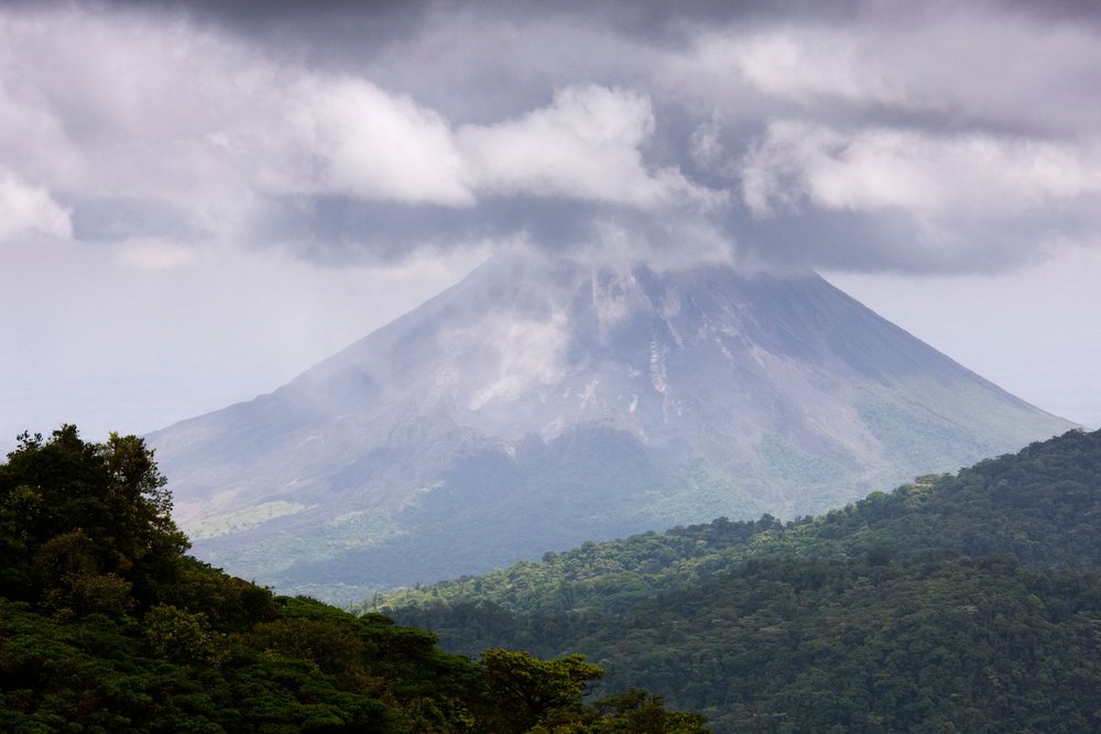 Arenal Volcano rises over green forested hills with clouds and mist near its summit under an overcast sky, Arenal Volcano National Park, Alajuela.