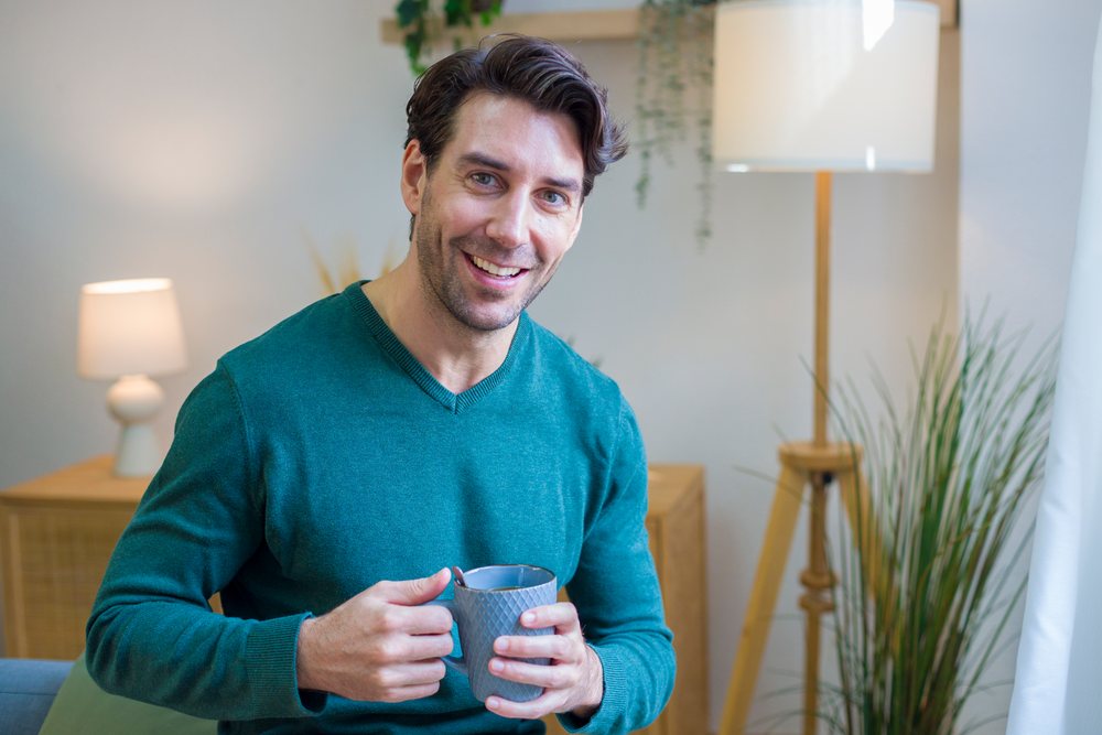 Man smiling while holding a cup of coffee in a cozy indoor home setting with modern lighting and plants, Technology, young adult.