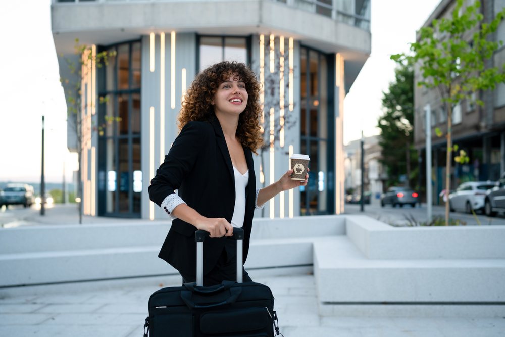 Business woman with curly hair walks on a sidewalk outside a modern building, holding a coffee cup and pulling a black suitcase, Madrid.