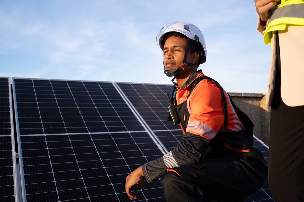 Worker wearing helmet and protective clothing crouches next to rooftop solar panel installation under a clear blue sky, Madrid, Spain.