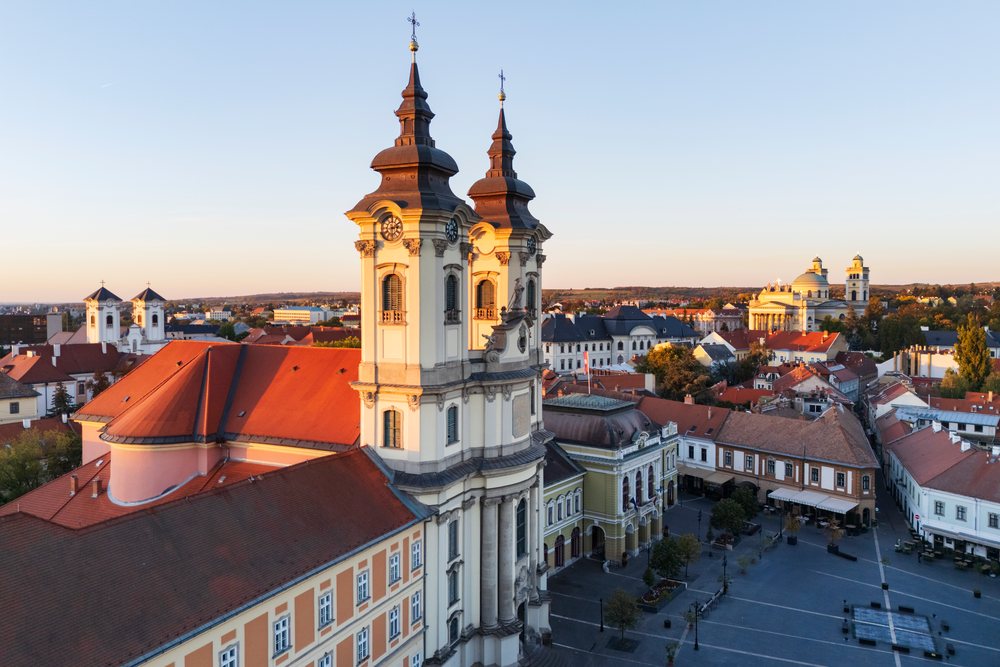 Church with twin towers and clock faces overlooking Dobó Square at Sunset in historic Eger, Hungary cityscape.