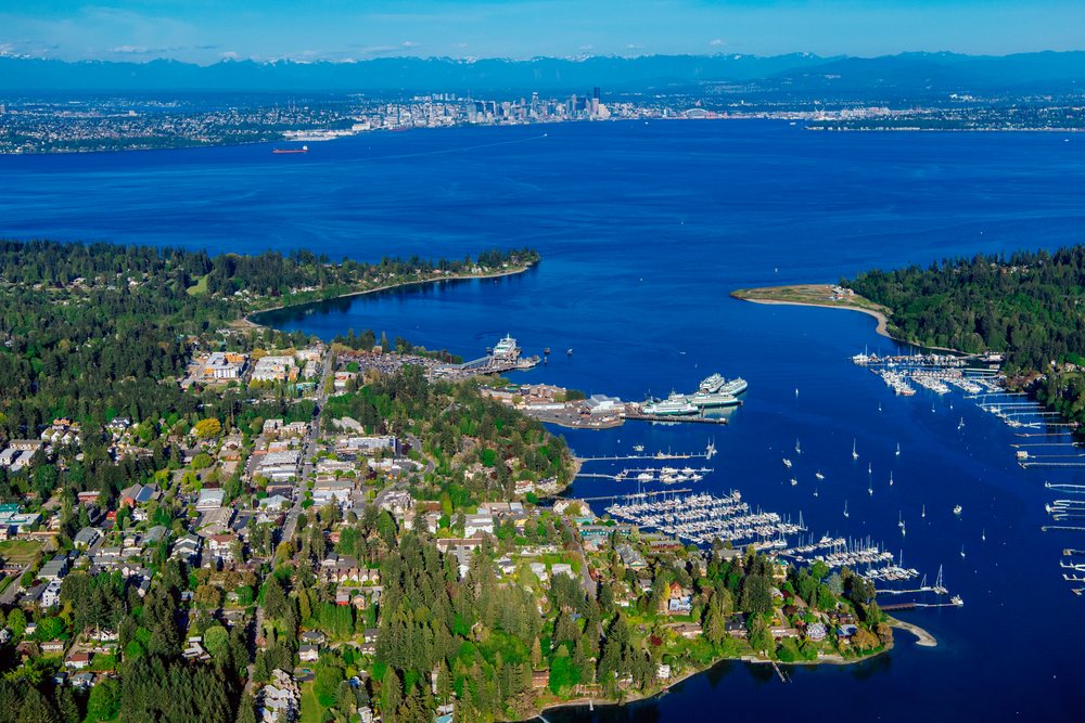 Aerial view shows boats and ferries docked at Eagle Harbor marina on Bainbridge Island with Puget Sound and Seattle skyline beyond, Salish Sea, USA.