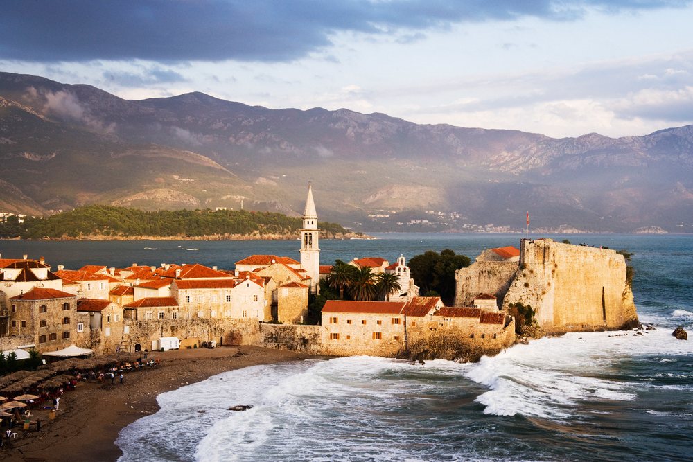 Historic coastal town with fortress and church tower overlooking the Adriatic Sea and mountain range.