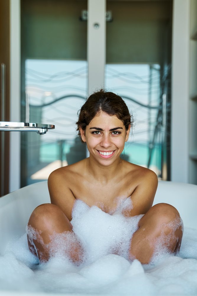 A young woman smiles while sitting in a bubble bath in a modern bathroom with a view in the, Maldives.