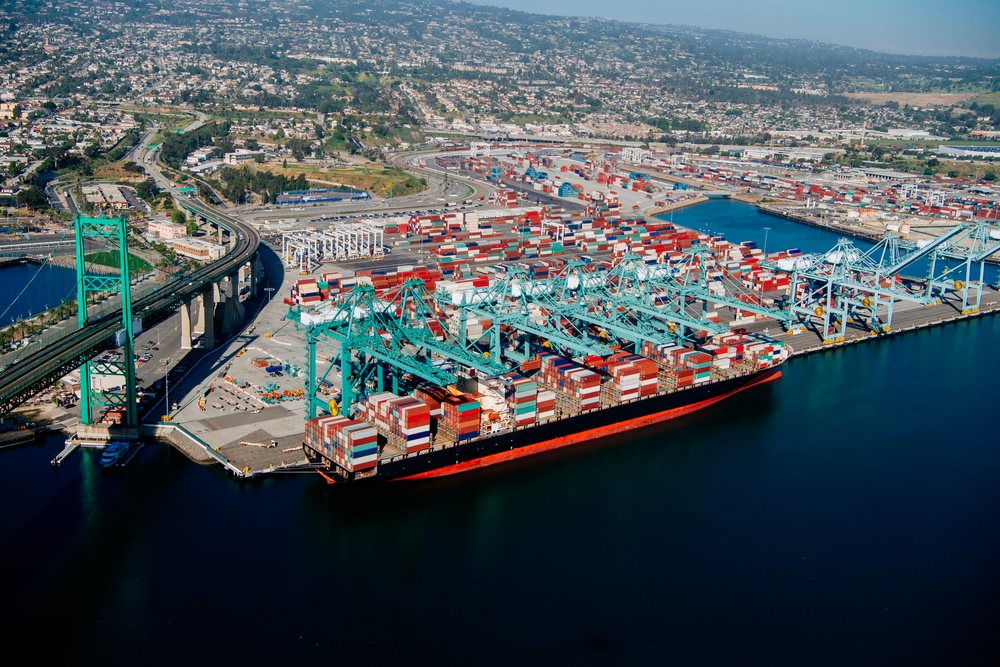 Container ship loaded with shipping containers docked at a major cargo port with cranes and a bridge visible in the background, Port of Long Beach, CA, USA.