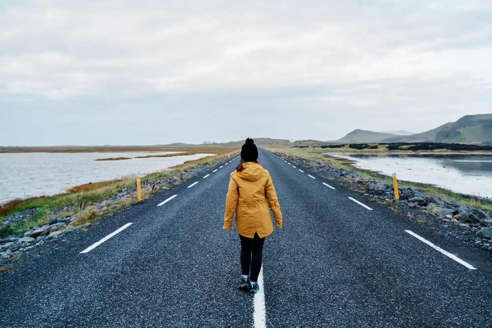 A white woman walks on an empty road surrounded by water and hills