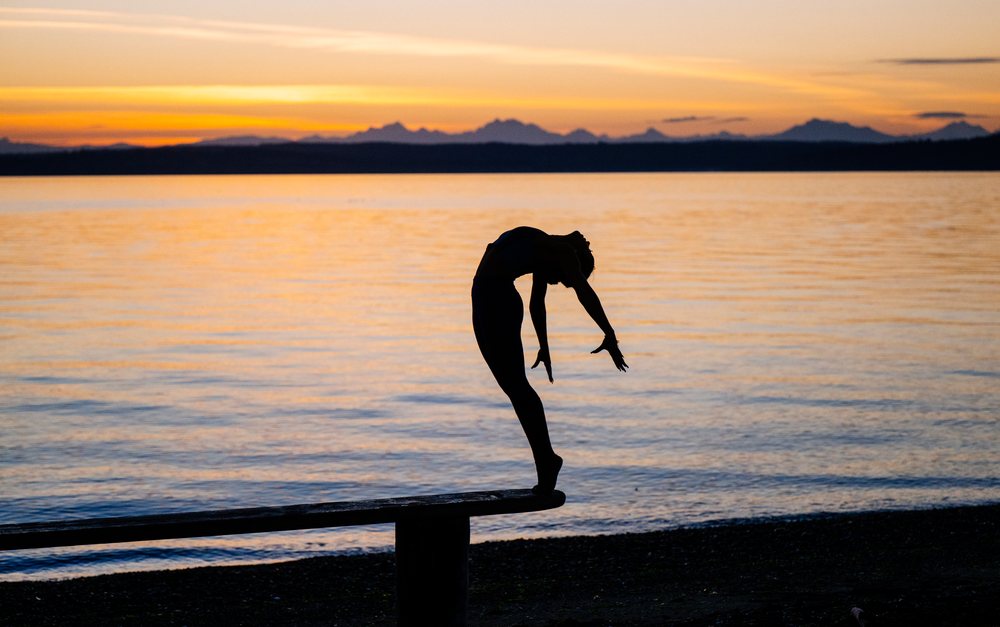 Woman performing an acrobatic yoga pose balancing on a bench at sunset over a calm lake, USA.