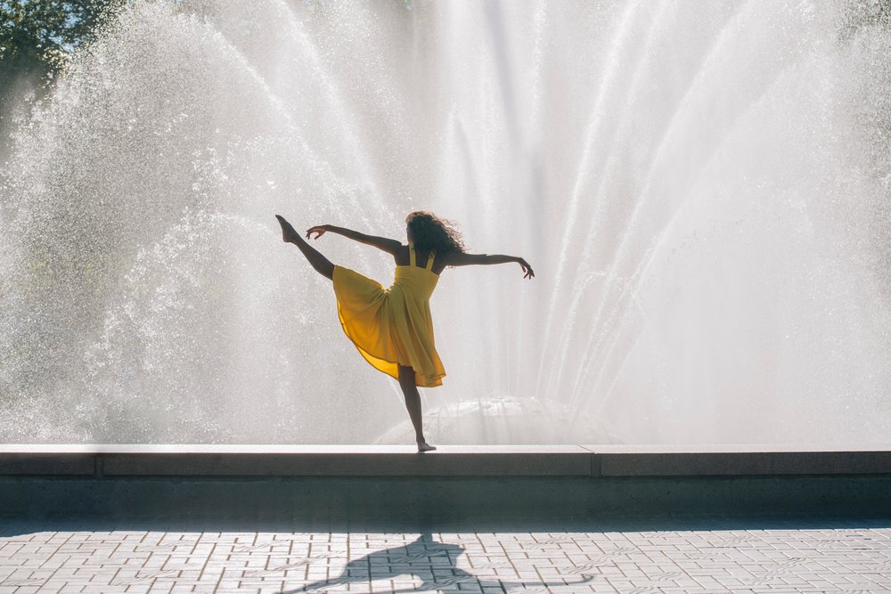 Female dancer in a yellow dress performs a balancing pose outdoors by a large water fountain, USA.