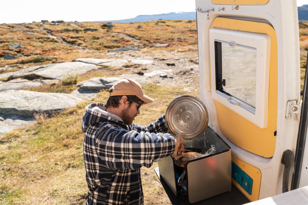 Man cooking meat on a portable stove outside a campervan in a rocky rural landscape in Norway during daylight.