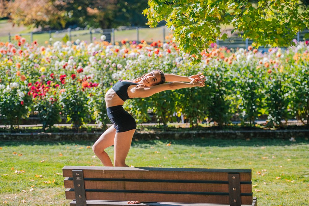 Woman contortionist performing a stretch on a bench in a sunny flower garden, Tacoma, WA, USA.