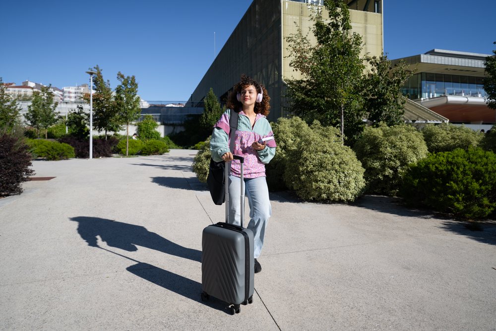 Woman walking outdoors on a sidewalk in an urban setting carrying a gray suitcase and wearing purple headphones and a backpack, Spain.