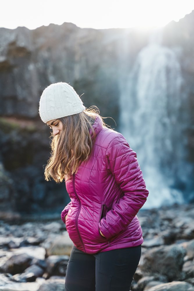 A woman in a purple jacket and white beanie walks on rocky terrain near a waterfall, Iceland.