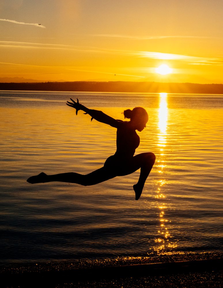 Silhouette of a young woman jumping in an acrobatic pose at sunset by the water, yoga, acrobatics, USA.