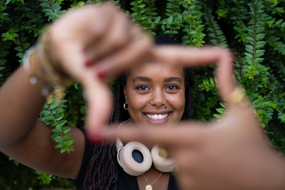 A smiling woman with braided hair frames her face with her hands outdoors against green foliage, urban, Santiago De Compostela, Spain.