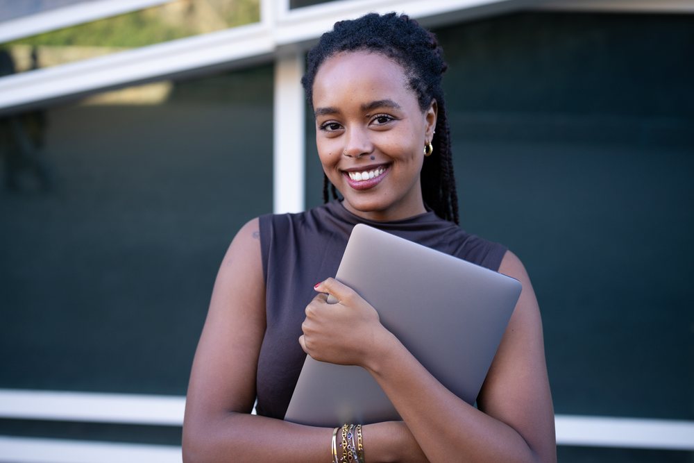 Woman smiling while holding a closed laptop outdoors in front of a dark building wall with white framing, Spain.