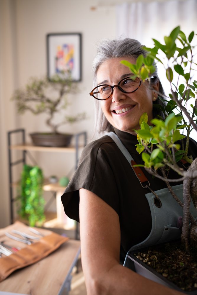Smiling woman with gray hair and glasses holds a bonsai plant indoors near gardening tools and shelving, Spain.