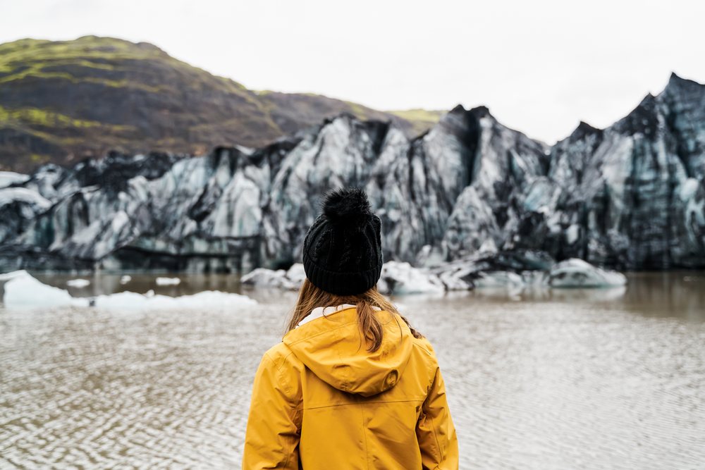 A white person wearing a yellow jacket looks out over a glacier lake