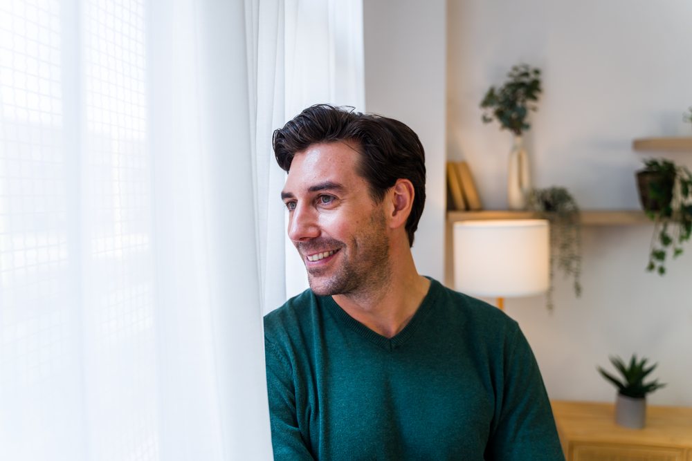 Man with facial hair wearing a green sweater smiles while looking out a window in a modern living room with plants and a lamp, Spain.