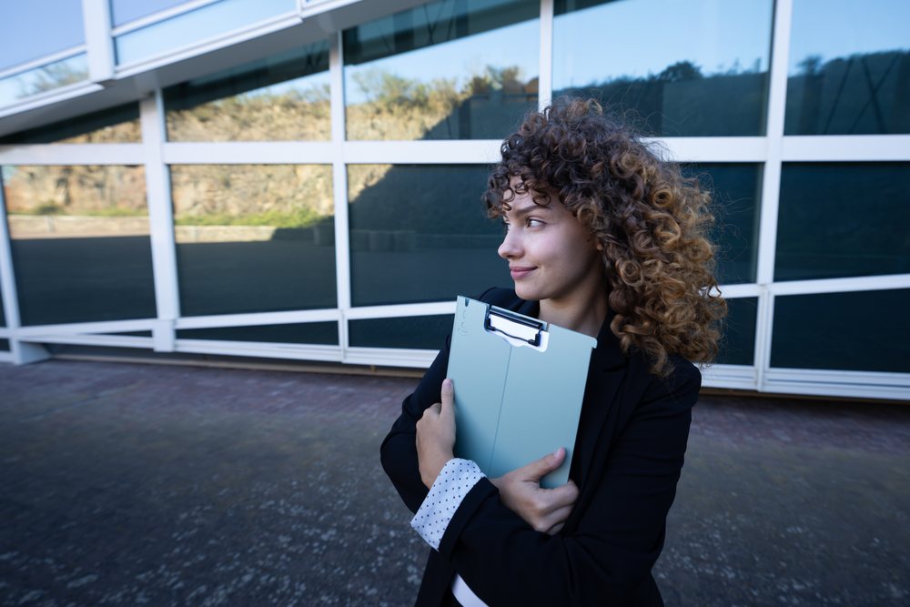 Businesswoman holding a folder and looking aside outside a modern office building, business trip, Spain, Santiago De Compostela.