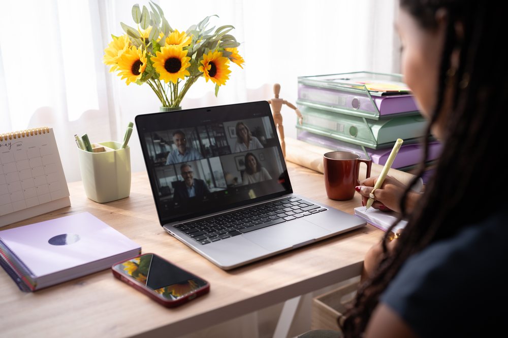 A woman writes in a notebook while attending a video call on her laptop in a bright home office, remote work, student, Spain.