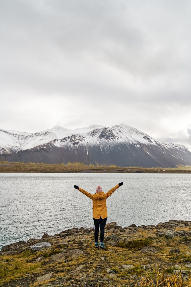 A person wearing a yellow jacket and pink hat stands with arms outstretched on a rocky shore by a lake with snow-covered mountains, Iceland.