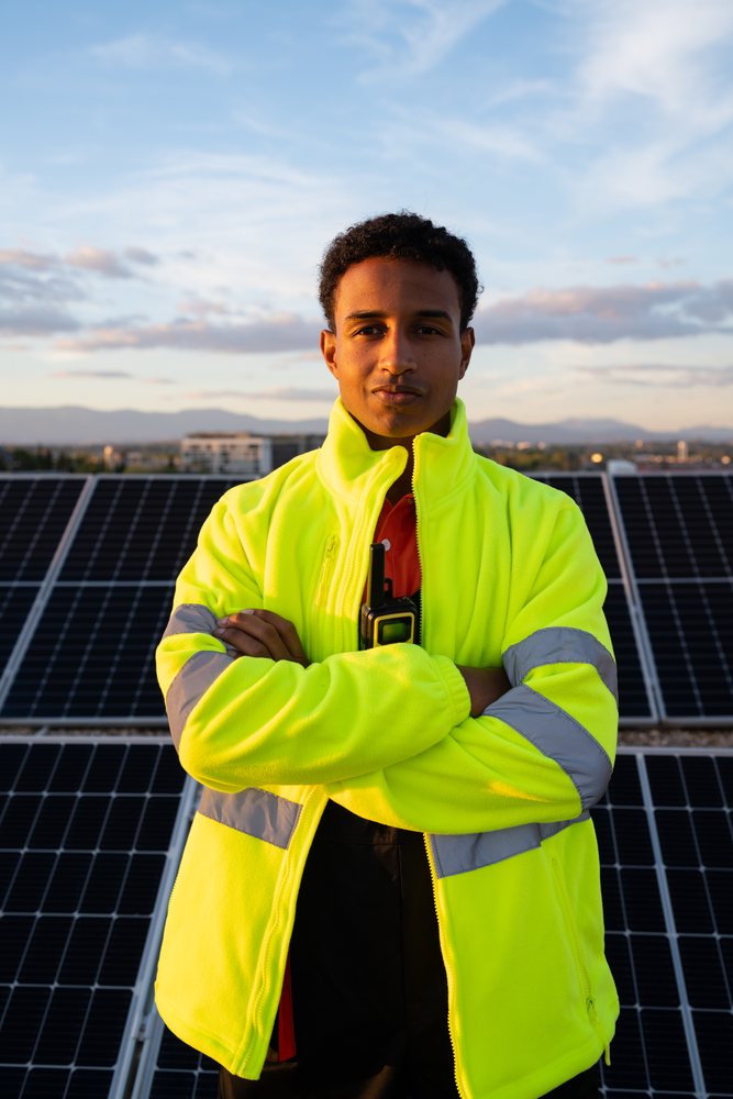 Man wearing a high visibility jacket stands outdoors by solar panels during sunset with a communication device clipped to his jacket, Madrid, Spain.