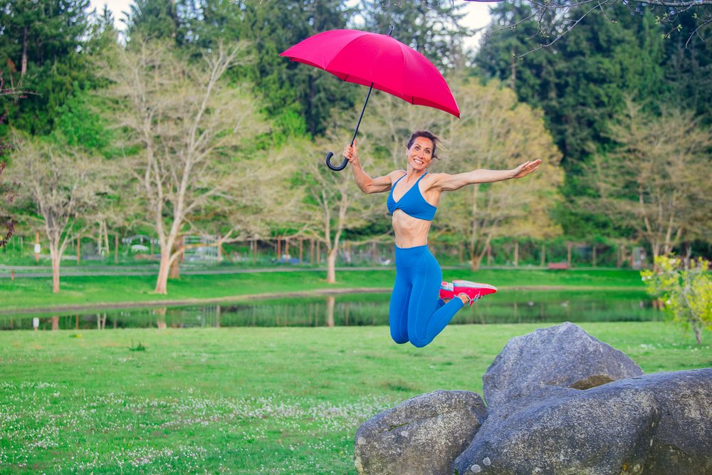 Smiling woman in blue sportswear jumps holding a red umbrella above rocks in a grassy park near a pond, Calisthenics, USA.