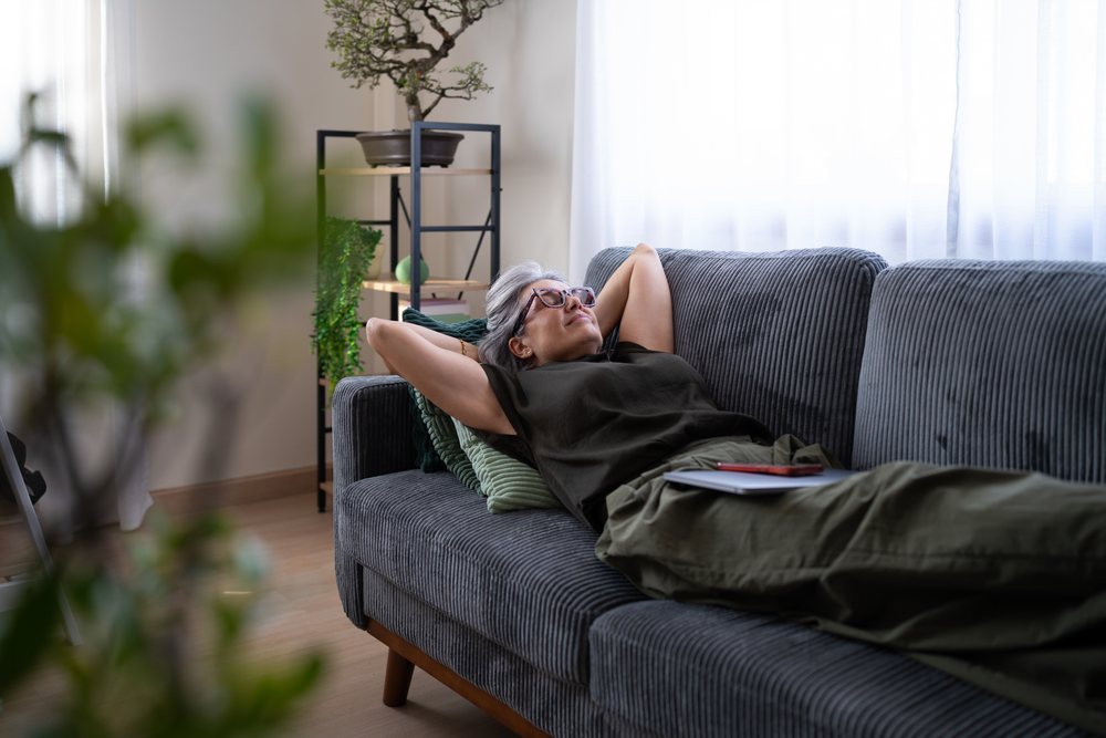 Mature woman with gray hair and glasses rests on a gray couch in a softly lit living room with green pillows and a wooden floor, Spain.