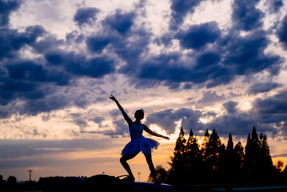 Ballet dancer in tutu performs a graceful silhouette pose outdoors against a dramatic cloudy sunset sky, USA.