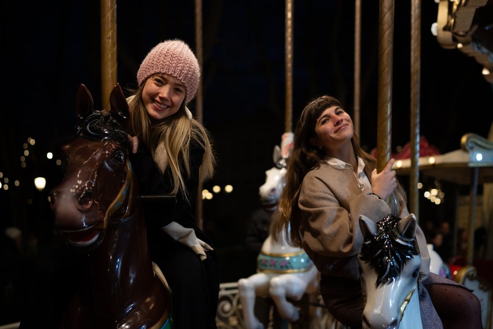 Two women smile while riding carousel horses at a Christmas market in Madrid at night, Spain.