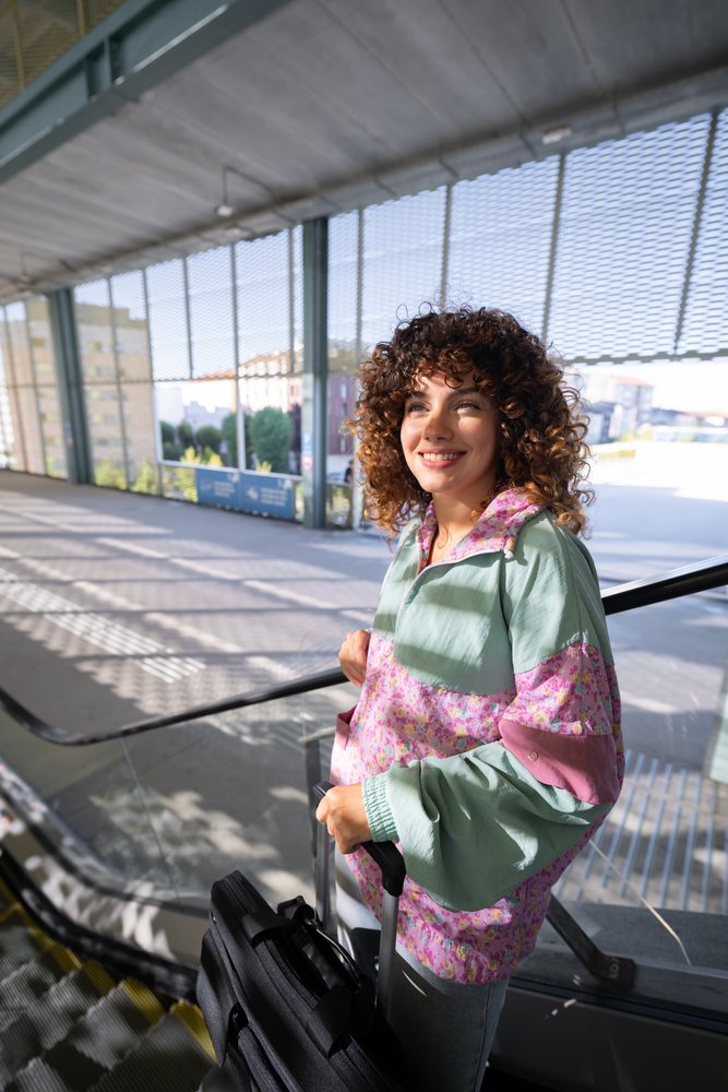 Smiling woman with curly hair holds luggage on an escalator in an urban outdoor setting during daytime, Spain.