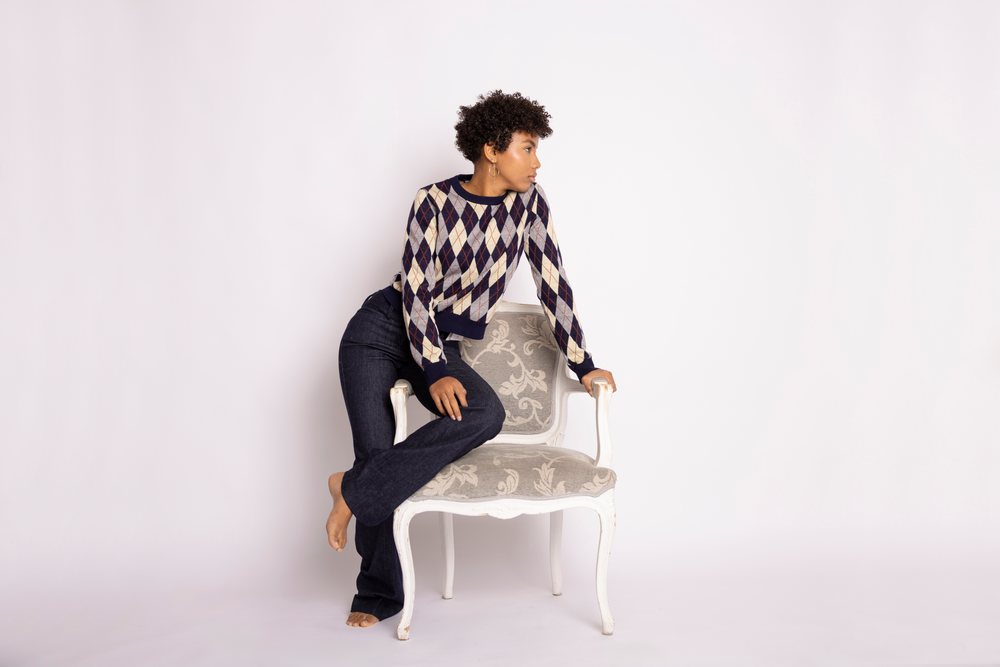 Woman with curly hair wearing an argyle sweater and jeans sits barefoot on a patterned chair in a studio with a white background, fashion, young culture.