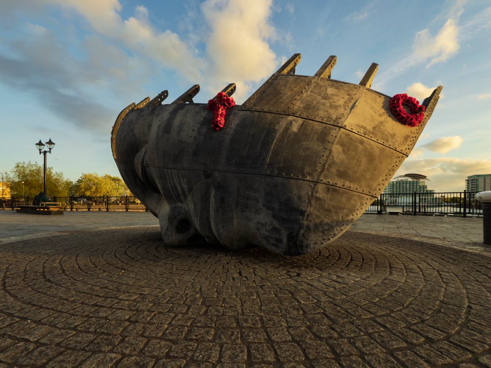 Large metal Ship's Prow sculpture with red memorial wreaths on circular cobblestone paving at Cardiff waterfront, Landscape, Glamorgan, Wales.